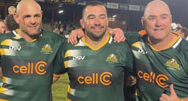 Three men in rugby jerseys stand smiling on a field. They link arms, showing camaraderie and joy. The stadium lights shine brightly in the background.