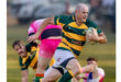 A rugby player in a green and yellow striped jersey runs with the ball, focused and determined. Two players in pink and blue jerseys chase him on a grassy field.
