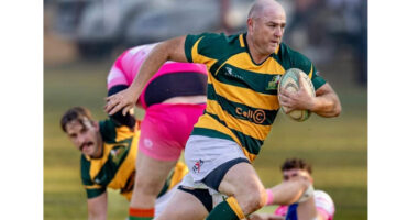 A rugby player in a green and yellow striped jersey runs with the ball, focused and determined. Two players in pink and blue jerseys chase him on a grassy field.