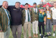 A group of seven smiling people stands outdoors. Four wear green and yellow striped blazers, one in a cap, two in sporty attire. Bright, cheerful mood.