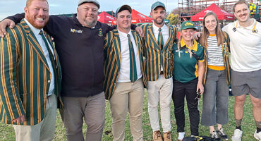 A group of seven smiling people stands outdoors. Four wear green and yellow striped blazers, one in a cap, two in sporty attire. Bright, cheerful mood.