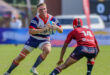 A rugby player in a white, blue, and red jersey holds a ball, facing a defender in red. The setting is a grassy field, with a focus on action and intensity.