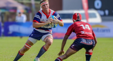 A rugby player in a white, blue, and red jersey holds a ball, facing a defender in red. The setting is a grassy field, with a focus on action and intensity.