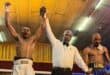 Boxer in white shorts, left, celebrates with arm raised by referee, center, while opponent in black shorts, right, stands defeated. Excited atmosphere.