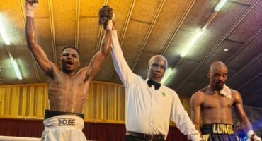Boxer in white shorts, left, celebrates with arm raised by referee, center, while opponent in black shorts, right, stands defeated. Excited atmosphere.