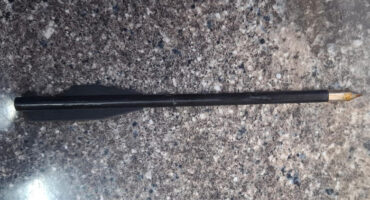 A black dart with a metal tip laying on a speckled gray stone surface. The dart is horizontally positioned, with minimal shadows.