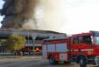 A fire truck is parked near a large building engulfed in flames and heavy black smoke. The scene is tense, highlighting emergency response efforts.