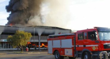 A fire truck is parked near a large building engulfed in flames and heavy black smoke. The scene is tense, highlighting emergency response efforts.