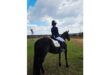 A person in an equestrian outfit sits on a black horse, facing a scenic outdoor horizon under a partly cloudy sky, conveying calm and focus.