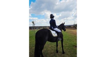 A person in an equestrian outfit sits on a black horse, facing a scenic outdoor horizon under a partly cloudy sky, conveying calm and focus.