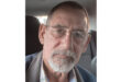 An older man with glasses and a white beard sits in a dimly lit car, gazing seriously at the camera. He wears a blue jacket and a checkered shirt.