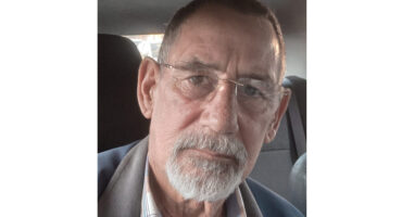 An older man with glasses and a white beard sits in a dimly lit car, gazing seriously at the camera. He wears a blue jacket and a checkered shirt.
