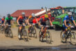 A group of cyclists in colorful jerseys race on a dirt path beside a red-roofed building and a green tractor, exuding energy and determination.