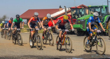 A group of cyclists in colorful jerseys race on a dirt path beside a red-roofed building and a green tractor, exuding energy and determination.