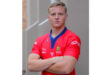 Young man in a red rugby jersey crosses his arms, exuding confidence. The background is a neutral-toned wall, creating a focused, serious tone.