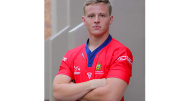 Young man in a red rugby jersey crosses his arms, exuding confidence. The background is a neutral-toned wall, creating a focused, serious tone.