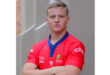 Young man in a red sports jersey, arms crossed, standing confidently against a gray urban backdrop. The tone is serious and determined.