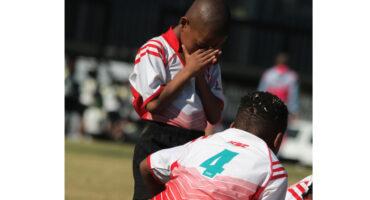 A young boy in a red and white sports jersey stands with hands covering his face, conveying emotion. Another child with a number 4 jersey is kneeling.