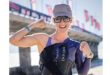 Woman in athletic gear smiles confidently, flexing her muscles with a bridge and flags in the blurry background. She wears a cap, glasses, and a watch.