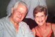 An elderly man and woman smiling, sitting close together. The man wears a striped shirt, and the woman, wearing a dress, conveys warmth and joy.