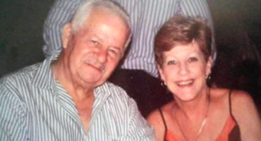 An elderly man and woman smiling, sitting close together. The man wears a striped shirt, and the woman, wearing a dress, conveys warmth and joy.