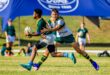 Two young rugby players in green and gold uniforms are in action on a grassy field. One is tackling the other, who is holding the ball tightly. A blue banner is visible in the background. The scene conveys energy and competition.