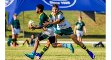 Two young rugby players in green and gold uniforms are in action on a grassy field. One is tackling the other, who is holding the ball tightly. A blue banner is visible in the background. The scene conveys energy and competition.