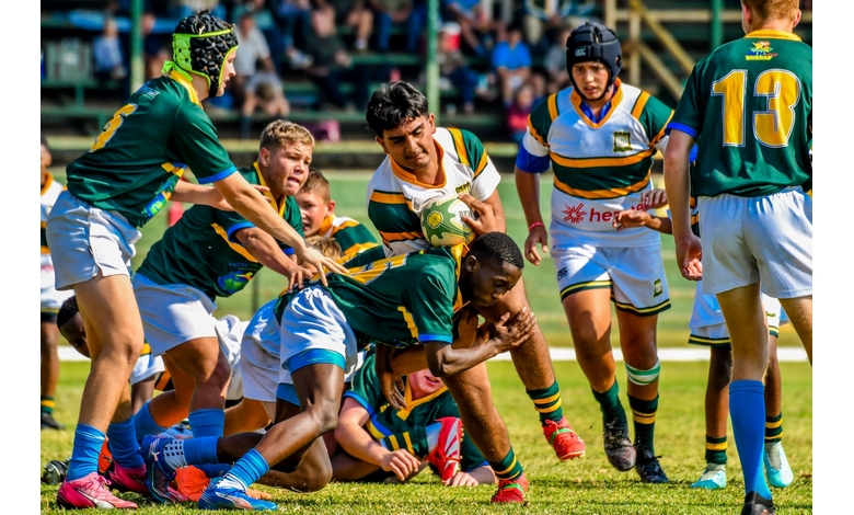 Players in green and blue uniforms engage in a dynamic rugby match, with one player tackling another holding a bright green ball, surrounded by teammates.