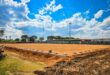 A sunny day at a sports field under construction, with a dirt surface and surrounding trees, light poles, and a distant building. The sky is blue with scattered clouds.