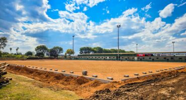 A sunny day at a sports field under construction, with a dirt surface and surrounding trees, light poles, and a distant building. The sky is blue with scattered clouds.