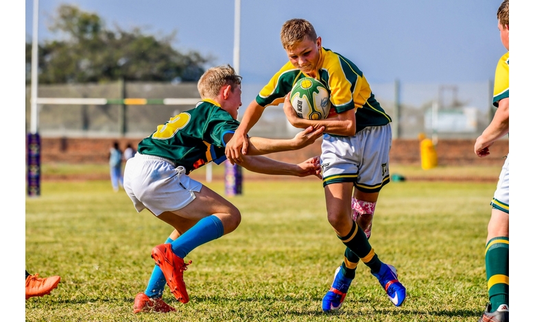 Two young rugby players in green and yellow jerseys compete on a sunny field. One carries the ball, evading a tackle from the other. Energetic scene.