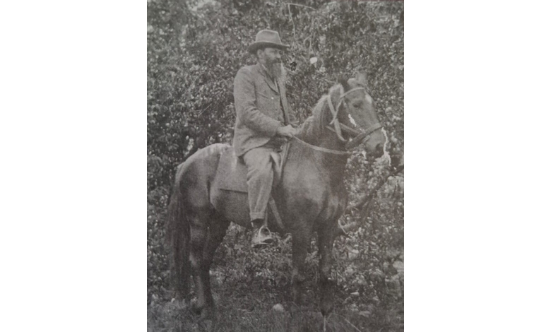 A black-and-white photo of a man in a hat and suit riding a horse. He is in a wooded area, exuding a calm, contemplative mood.