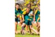 A group of young boys in green and yellow rugby uniforms playing rugby outdoors. One boy, focused, runs barefoot holding the ball. Bright and energetic scene.