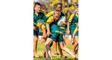 A group of young boys in green and yellow rugby uniforms playing rugby outdoors. One boy, focused, runs barefoot holding the ball. Bright and energetic scene.