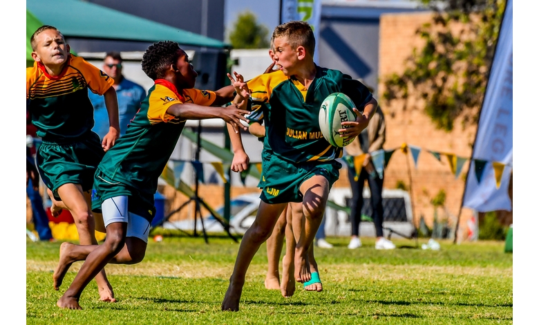 Two young rugby players in green uniforms compete during a sunny match. One, holding the ball, attempts to evade an opponent reaching to tackle him.