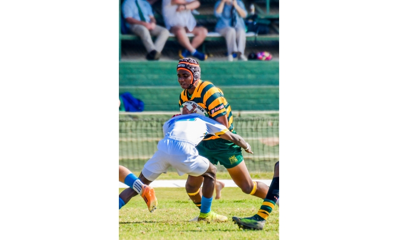 A rugby player in a striped jersey attempts to tackle an opponent wearing white. The action is intense, with spectators watching in the background.