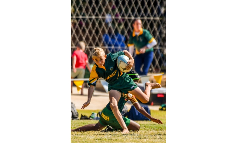 A young rugby player in a green and yellow jersey leaps skillfully over a defender on a grass field. Spectators are visible in the blurred background.