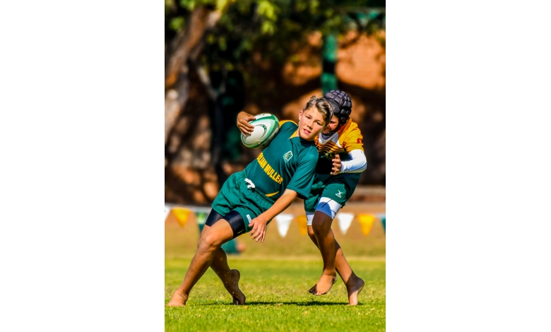 Two boys play rugby on a grassy field. One in a green jersey holds the ball, while the other in a yellow jersey attempts a tackle. Focused and dynamic.