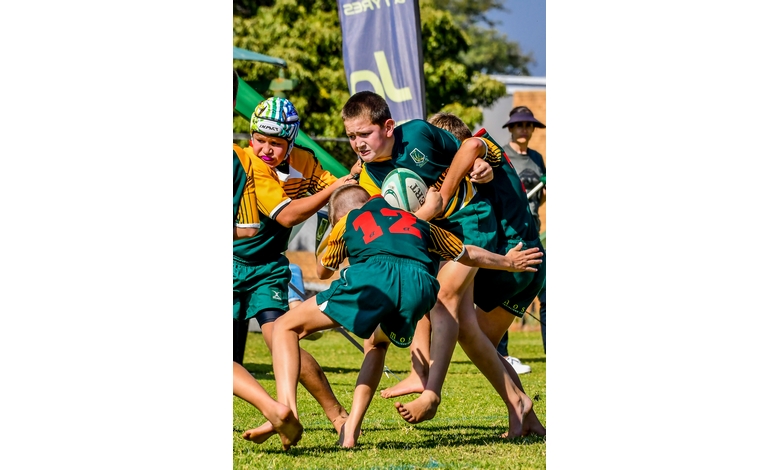 Youth rugby match with players in green and yellow tackling for the ball. Intense action and focus, with bright sunlight on the grassy field.