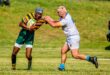A rugby player in a green and yellow striped jersey holds the ball, fending off an opponent in white. Action takes place on a grassy field.