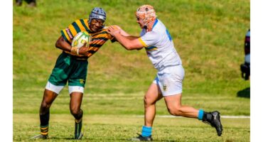 A rugby player in a green and yellow striped jersey holds the ball, fending off an opponent in white. Action takes place on a grassy field.