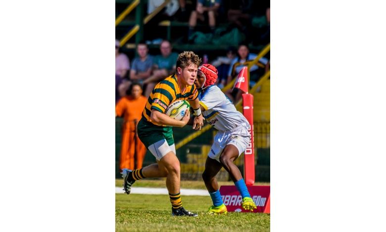A rugby player in a striped green and yellow jersey grips the ball tightly while evading a tackler. Intense focus and action on the field.
