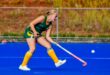A hockey player in a green and yellow uniform is hitting a ball on a bright blue field. The scene conveys focus and athletic action.