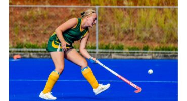 A hockey player in a green and yellow uniform is hitting a ball on a bright blue field. The scene conveys focus and athletic action.