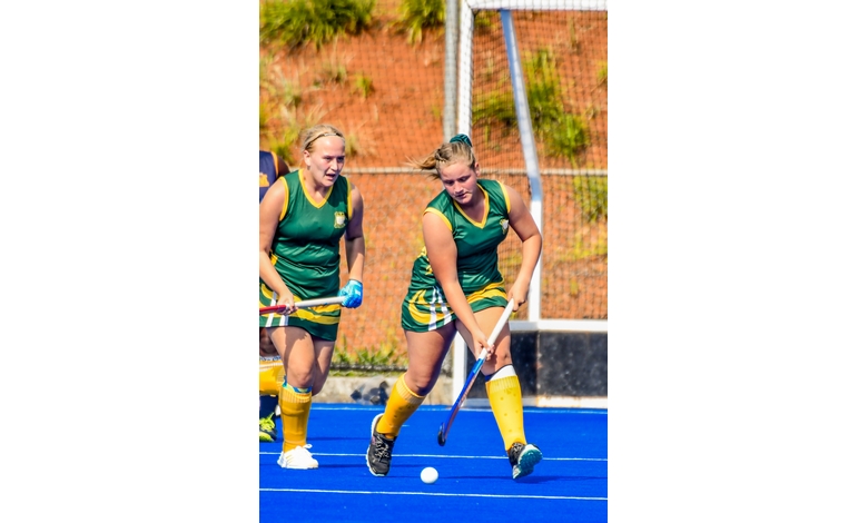 Two female field hockey players in green and yellow uniforms compete on a bright blue turf. One advances the ball with focus, while the other follows closely.