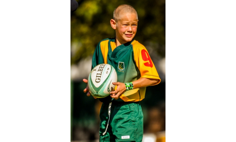 A young boy in a yellow and green rugby uniform holds a rugby ball on a sunny day. He looks determined, with trees blurred in the background.