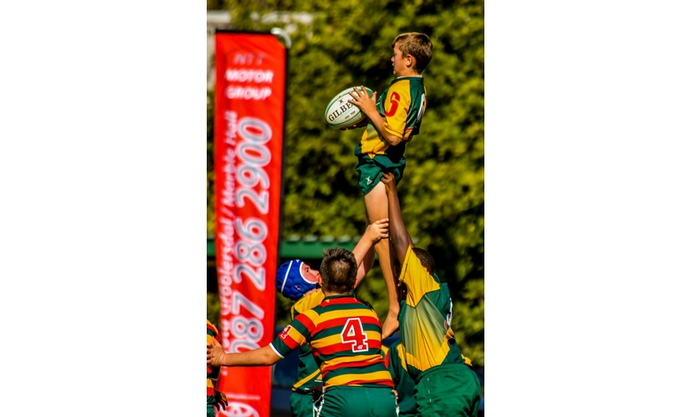 Young rugby player in yellow and green uniform is lifted by teammates, grasping a rugby ball. Red banner in background, lush greenery surrounding. Energetic scene.