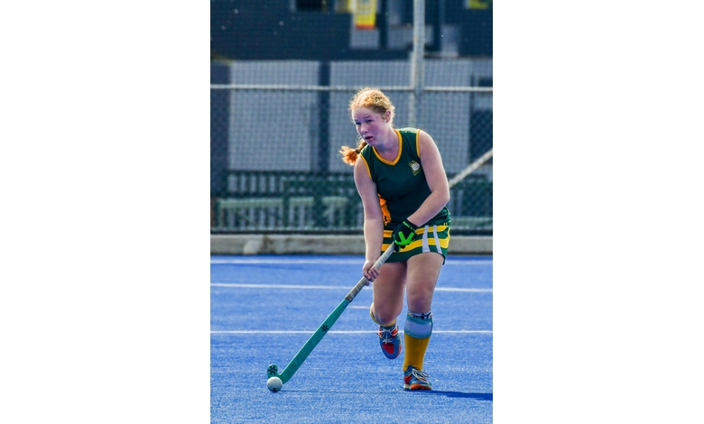 A field hockey player in green and yellow uniform focuses intently while dribbling a ball on a vibrant blue turf field, conveying determination.
