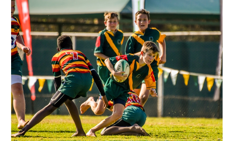 A youth rugby player in a green and yellow jersey charges forward clutching the ball, dodging two opponents in red and green. Focused action, teammates watch.