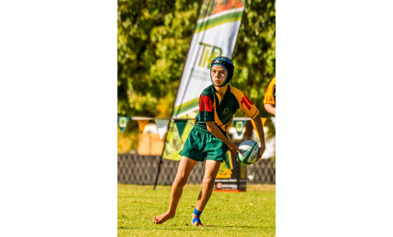 A young rugby player in green and gold gear, wearing a helmet, clutches a ball while running on a grassy field. A focused expression and a branded flag in the background suggest a competitive game atmosphere.